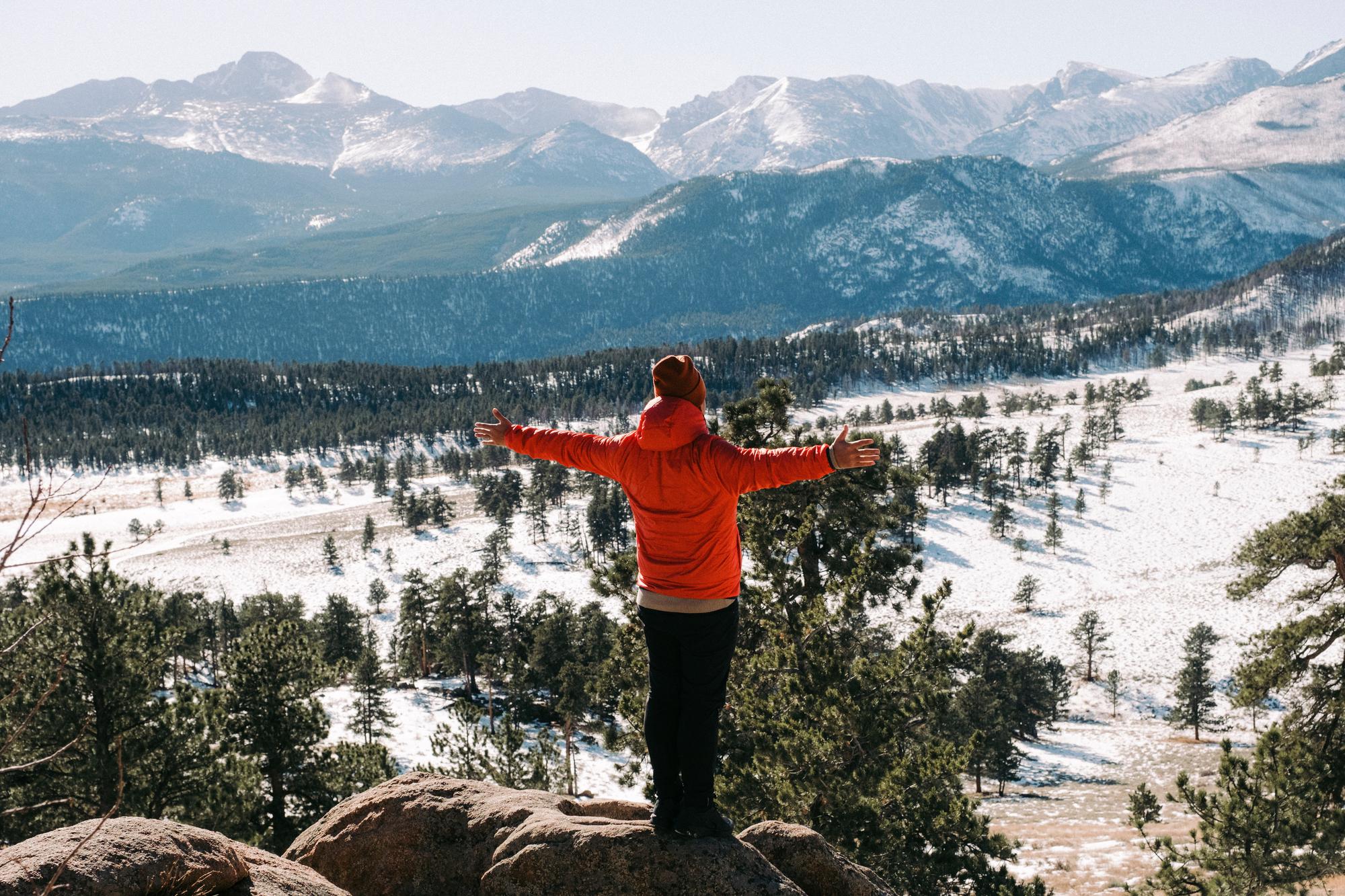 A figure standing with their back to us in a red winter jacket and hat, arms out stretched, atop a rocky area looking down into a valley surrounded by mountains - North Park, Jackson County Colorado