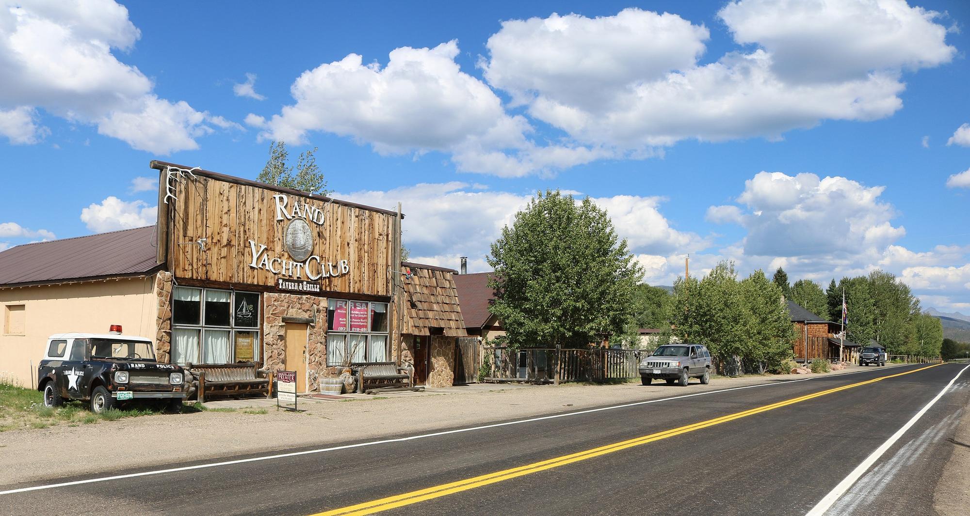 An exterior of a building with a sign that reads Rand Yacht Club Tavern & Grille - a for sale sign is visible in the window. An old fashioned police car is parked next to it. A highway stretches in the foreground - North Park, Jackson County Colorado