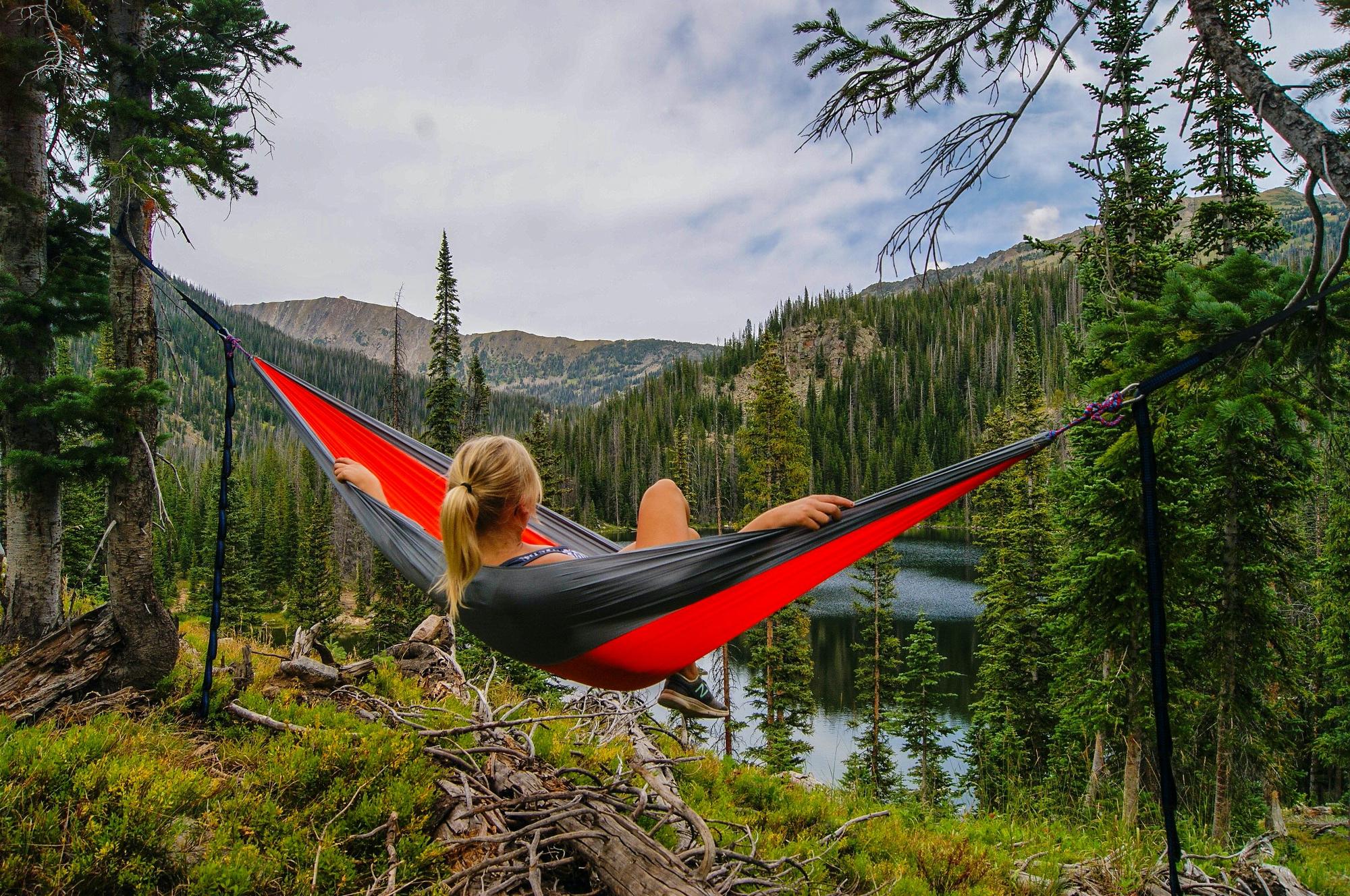 A young blonde woman with her back to us relaxes in a red hammock above a serene lake in a thickly wooded area. The valley is made up of thick tall trees - North Park, Jackson County Colorado