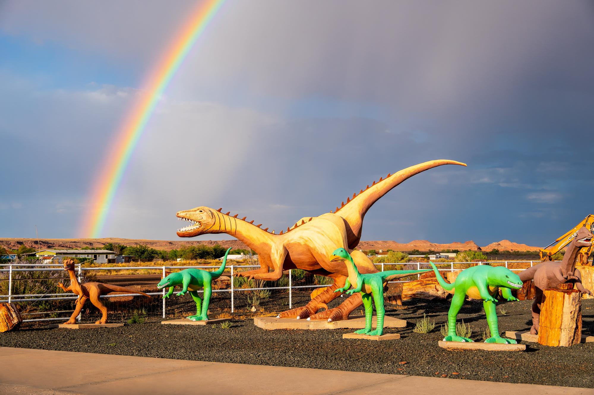 6 dinosaur statues in front of desert mountains with a rainbow in the sky