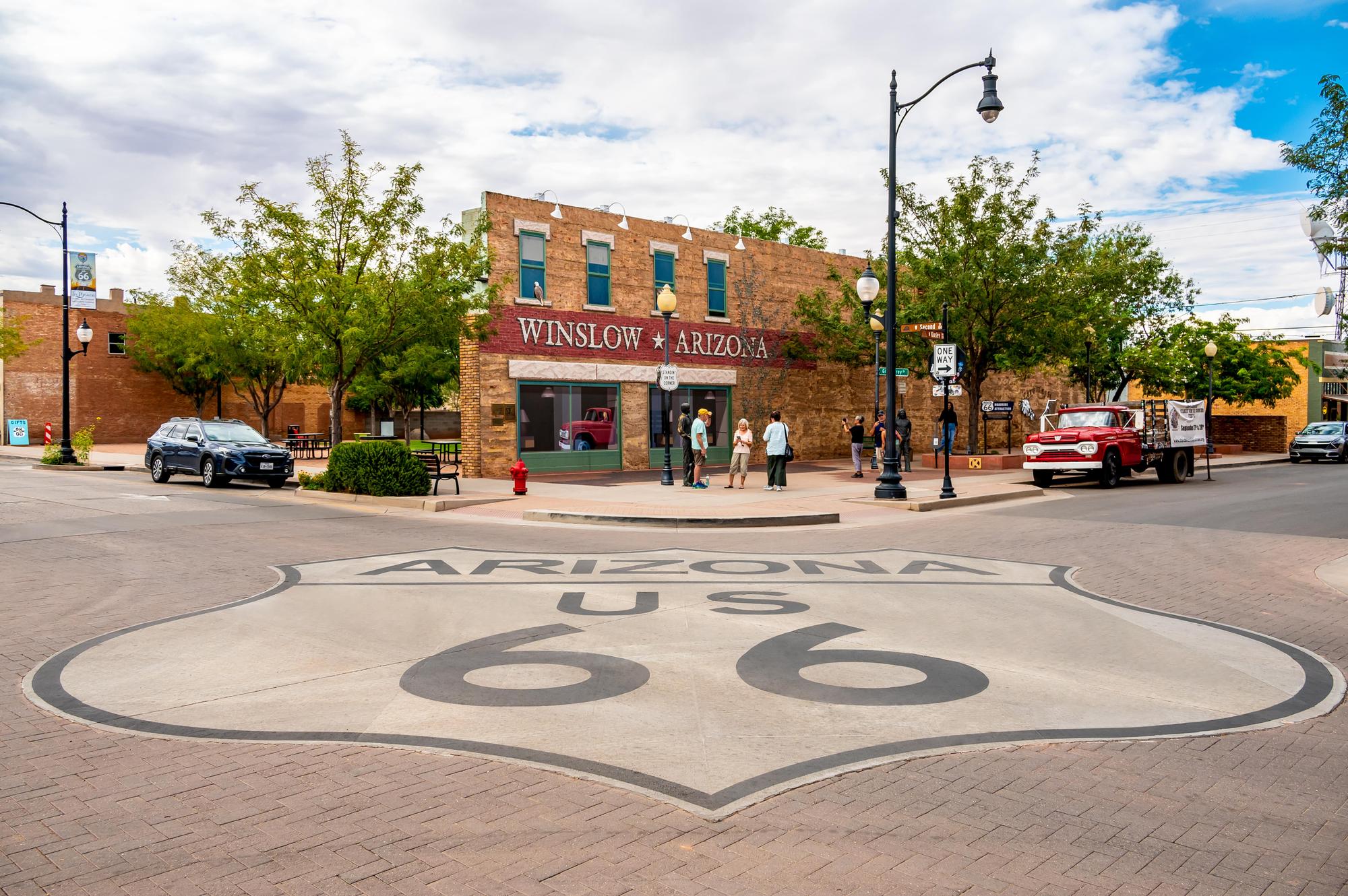Route 66 shield in the road at the intersection of the Standing on the Corner Park in Winslow, Arizona. Visitors taking photos with a bronze statue of Glenn Frey