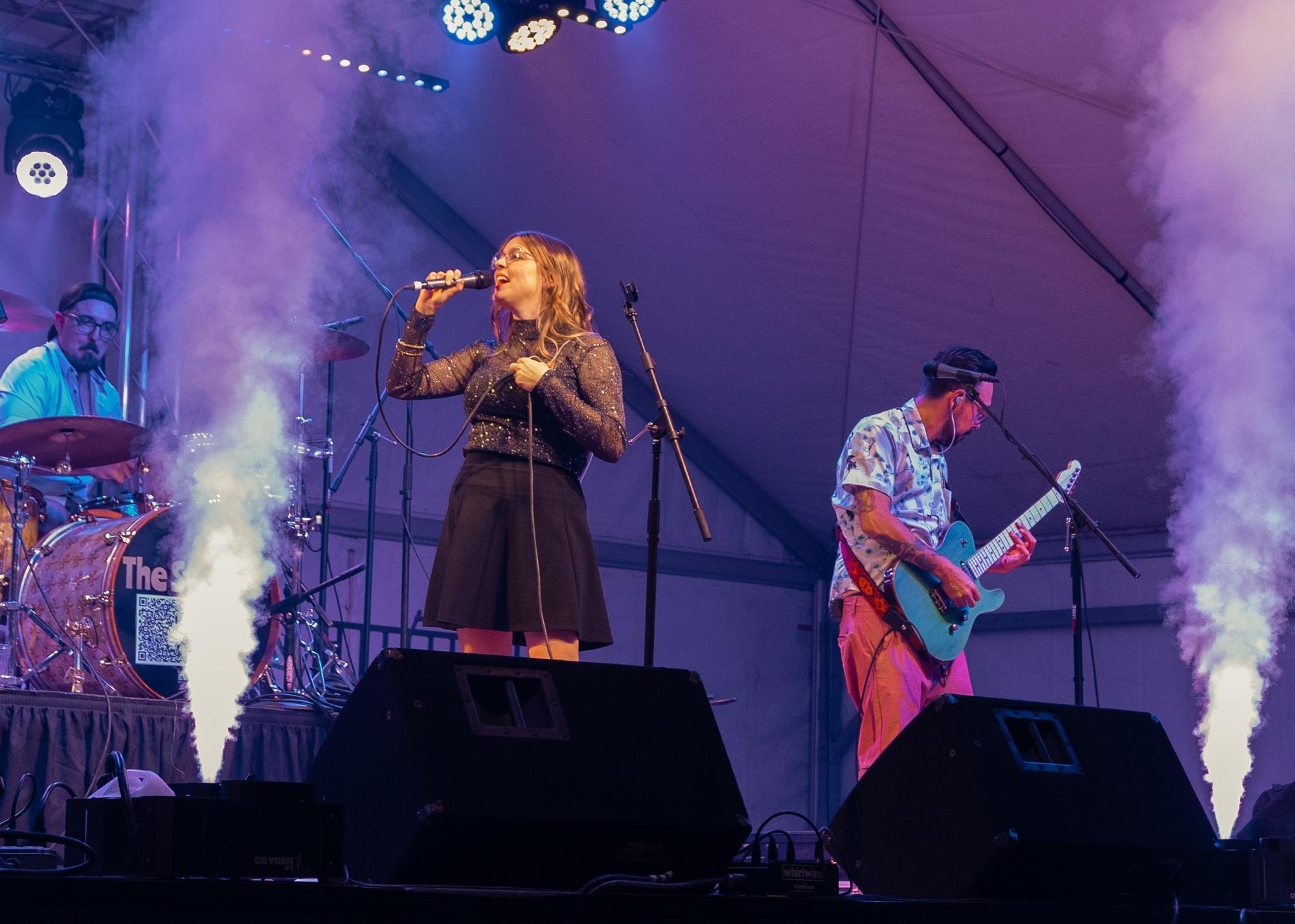 3 band members onstage, one man plays the drums, another plays the guitar, while a woman wearing a black sparkly dress is singing. 