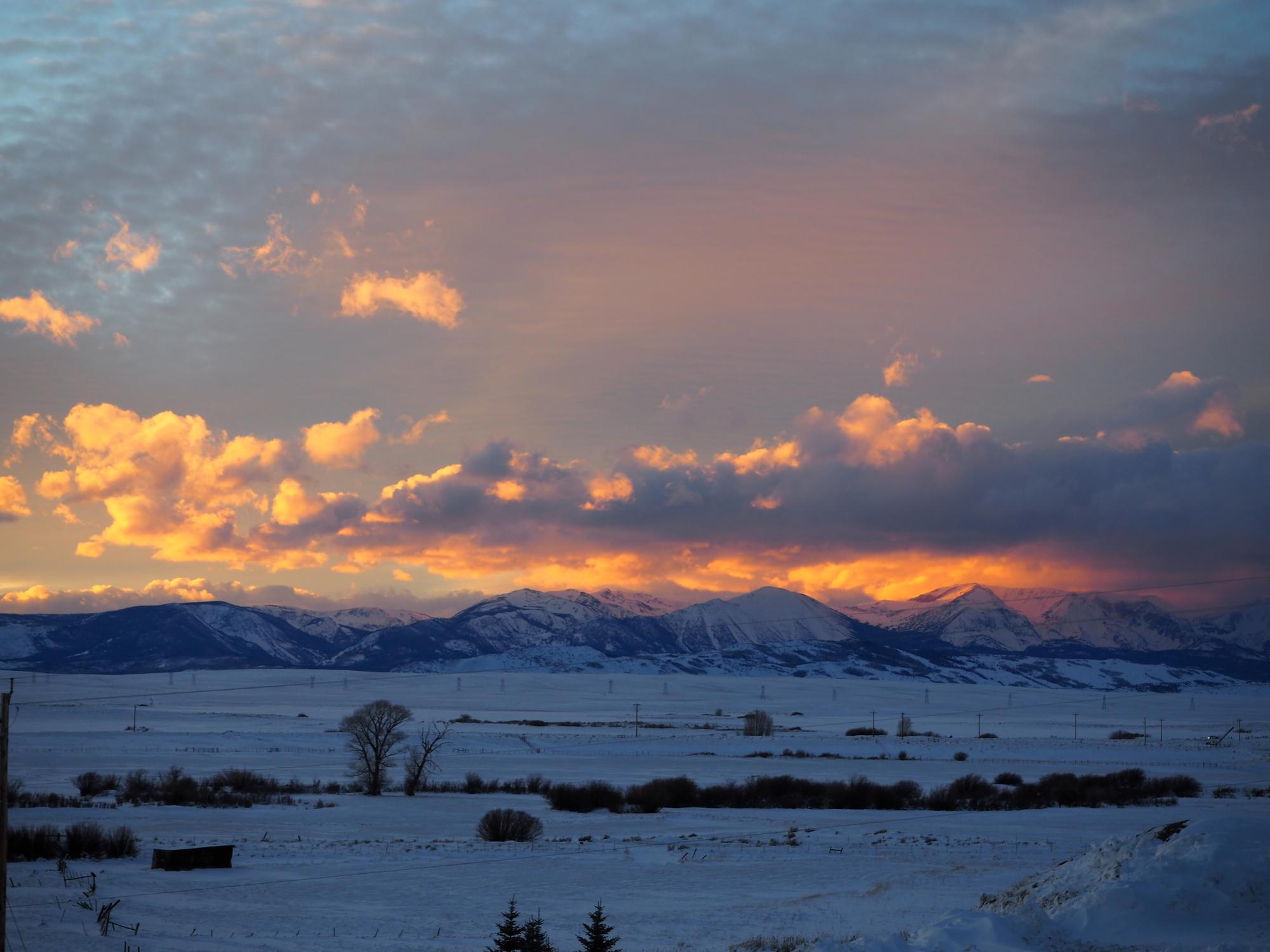A scenic landscape features snow-covered fields and shrubs in the foreground, leading to a distant mountain range. The sky is filled with clouds illuminated by the warm hues of a setting sun.