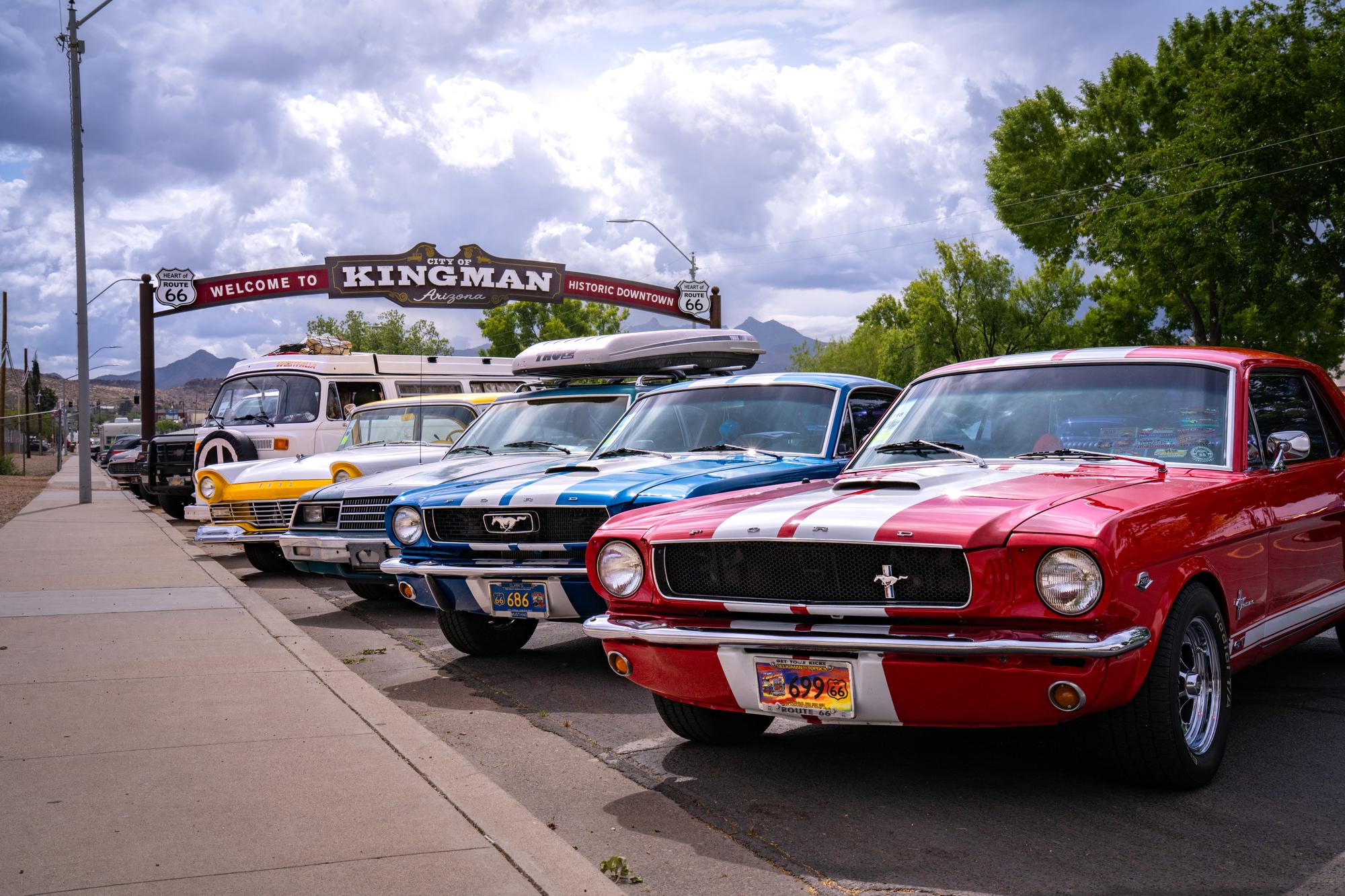 Vibrant classic cars parked along a sidewalk with the Welcome to Kingman arch in the background. 